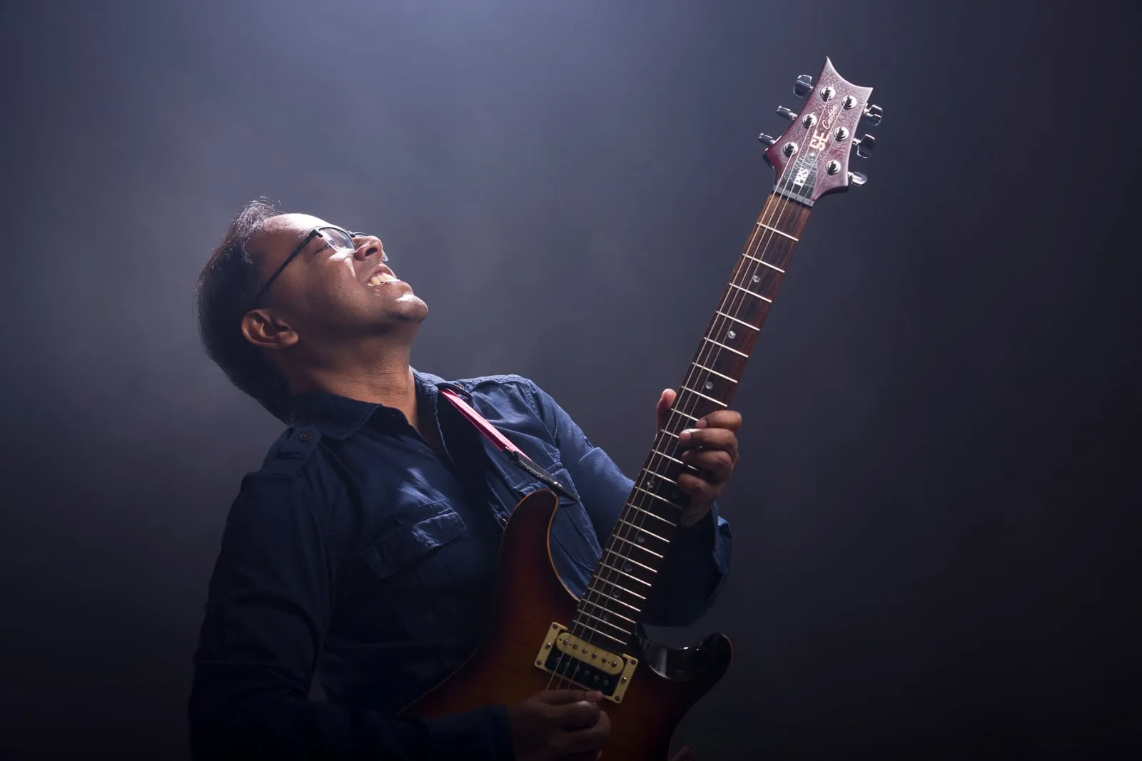 Bijoy Thangaraj playing an electric guitar under a spotlight, head tilted back mid-phrase